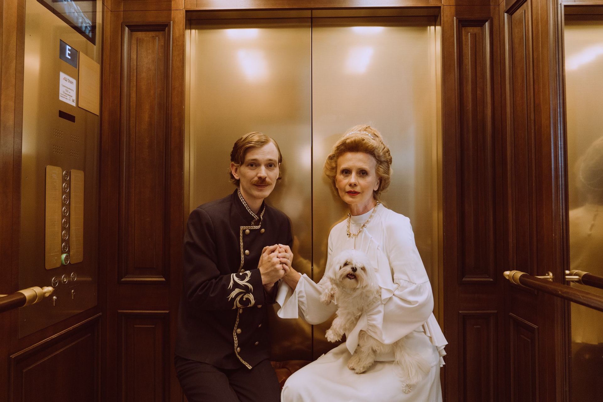 A hotel porter in an embroidered uniform beside a guest holding a small white dog, in a wood-panelled lift